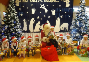 Chłopiec siedzi na kolanach Mikołaja, ogląda prezent. Wokół siedzi grupa dzieci.